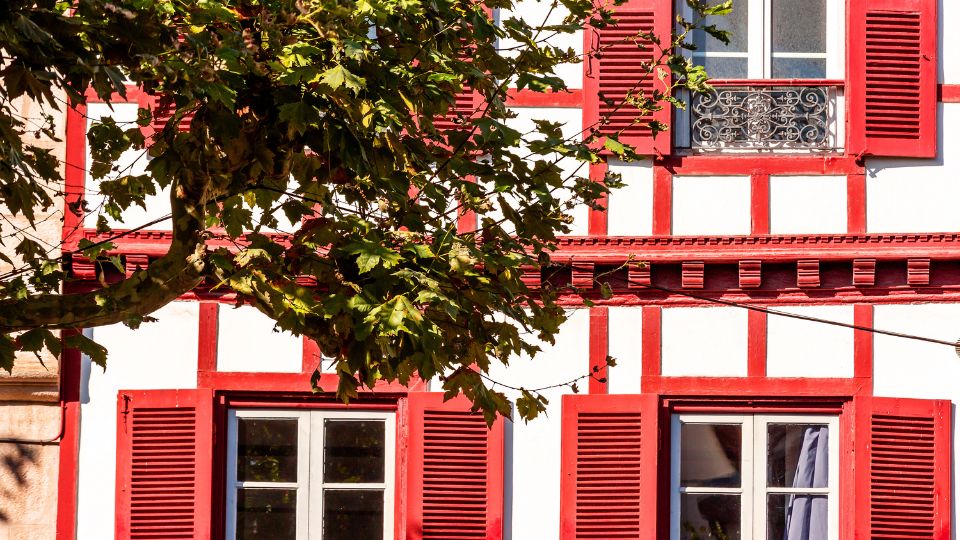 Maison traditionnelle basque Détail d'une maison traditionnelle basque à Anglet avec des volets rouges et des éléments architecturaux en bois rouge et blanc, partiellement ombragée par un arbre.
